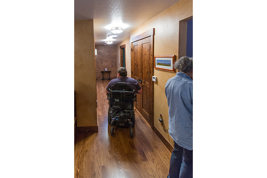 A man in a wheelchair travels down a spacious, stained wood floor hallway past a reachable thermostat control and lever-handled double doors while his wife fiddles with something just out of the picture.