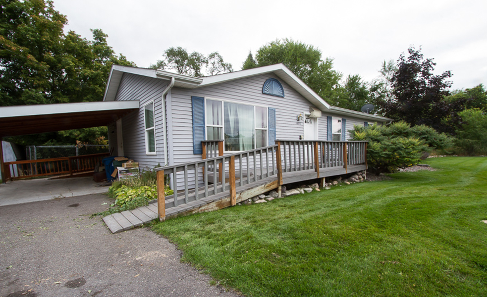 A grey, modular home features two long ramps connecting the front and back decks to a driveway.  The front deck is made out of grey composite boards, while the rear deck and ramp are made from brown stained wood boards.
