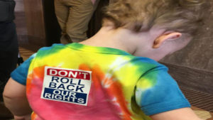 Young child wearing a tie-dye shirt with a "Don't Roll Back Our Rights" sticker on the back.