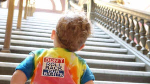 Young child looking up the Capitol Rotunda staircase. Child is wearing a tie-dye shirt with a "Don't Roll Back Our Rights" sticker on the back.