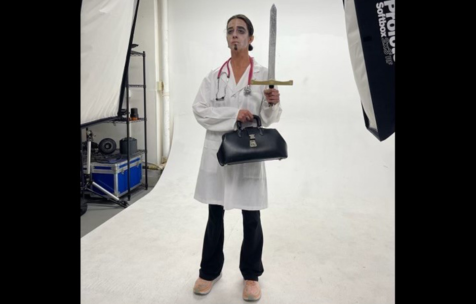 Heidi dresses up as a male doctor holding a sword and medical bag, while sporting a dark beard and white coat