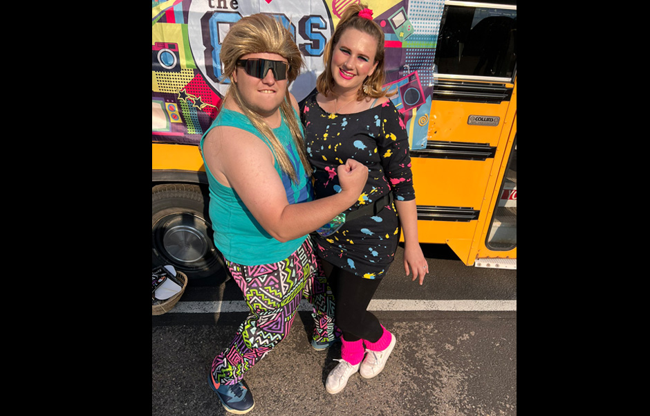 Tyler, dressed in a blonde mullet wig and brightly patterned pants, poses with Madison, who is dressed up in pink leg warmers and a black dress with bright paint splashes, in front of the short bus photo wall