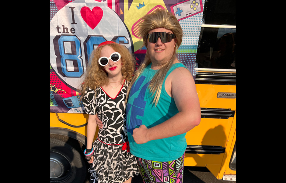 Molly, dressed in an authentic black and white 90's prom dress and white glasses, poses with Tyler, wearing a blonde mullet wig