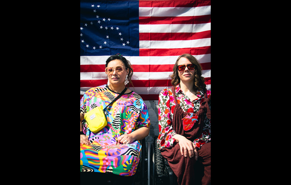 Two women wear fashionable clothing from Betty's while posing in front of the American flag with the stars arranged into an image of a person in a wheelchair