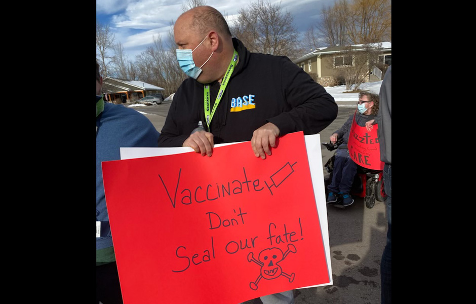 Jason holds a sign saying "Vaccinate don't seal our fate!"
