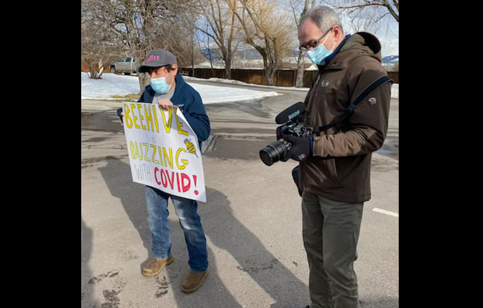 Mike stands with a sign stating "Beehive is Buzzing with COVID" and an individual from the Missoulian prepares a cameras to take pictures