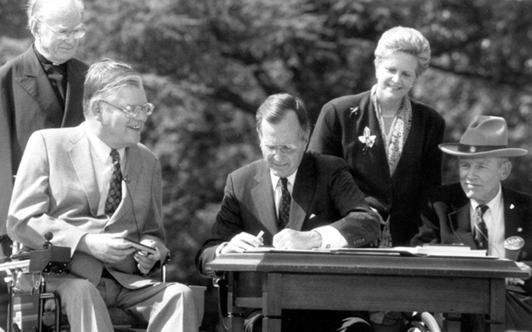 In this photograph taken on July 26, 1990, President George H. W. Bush signs the Americans with the Disabilities Act during a ceremony in the Rose Garden. Sitting beside him from left to right are Evan Kemp, Chairman of the Equal Employment Opportunity Commission, and Justin Dart, Chairman of the President’s Committee on Employment of People with Disabilities. Standing behind him from left to right are Reverend Harold Wilke and Swift Parrino, Chairperson, National Council on Disability.
