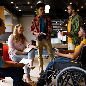 A group of 5 peers talking in a circle conversing, one is a wheelchair user, two people are sitting down, and two people are standing