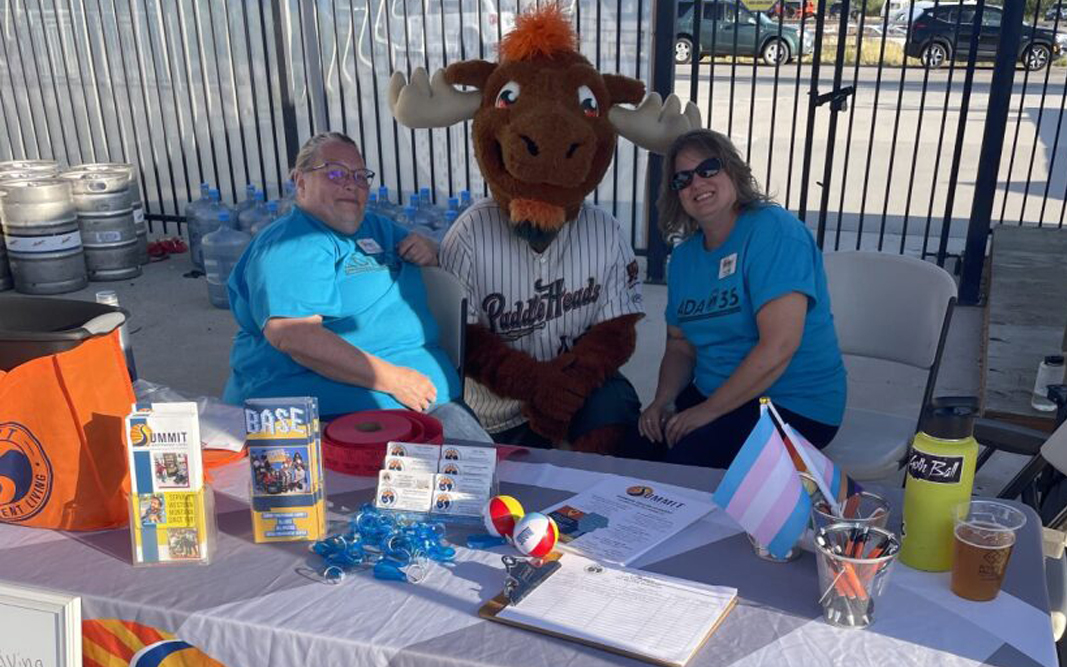 Two Summit staff members hanging out with the Missoula Paddle Head's mascot behind a Summit informational table