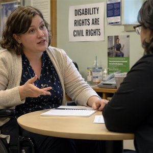 Woman using a wheelchair speaks with another person at a round table; a “Disability Rights Are Civil Rights” sign hangs behind them.
